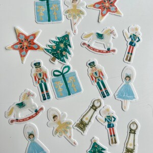 Könnte beinhalten: Eine Sammlung von Aufklebern zum Thema Weihnachten mit verschiedenen Designs. Die Aufkleber zeigen Geschenkboxen, Ballerinas, Sterne, Weihnachtsbäume, Schaukelpferde, Nussknacker, eine Maus und Standuhren. Die Aufkleber sind in Blau, Grün, Rot, Gelb und Weiß gehalten.