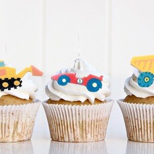 May include: Three cupcakes decorated with construction vehicle themed toppers. The cupcakes are frosted with white frosting and have a yellow, red, and orange construction vehicle on top.