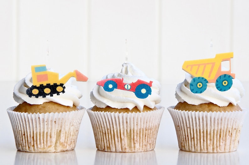 May include: Three cupcakes decorated with construction vehicle themed toppers. The cupcakes are frosted with white frosting and have a yellow, red, and orange construction vehicle on top.