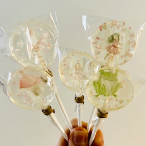 Könnte beinhalten: Fünf transparente Lollis mit skurrilen Designs, darunter Feen und ein Frosch, sind einzeln verpackt. Die Lollis haben weiße Stiele und sind mit rosa und weißen Akzenten verziert. Ein süßer Leckerbissen.