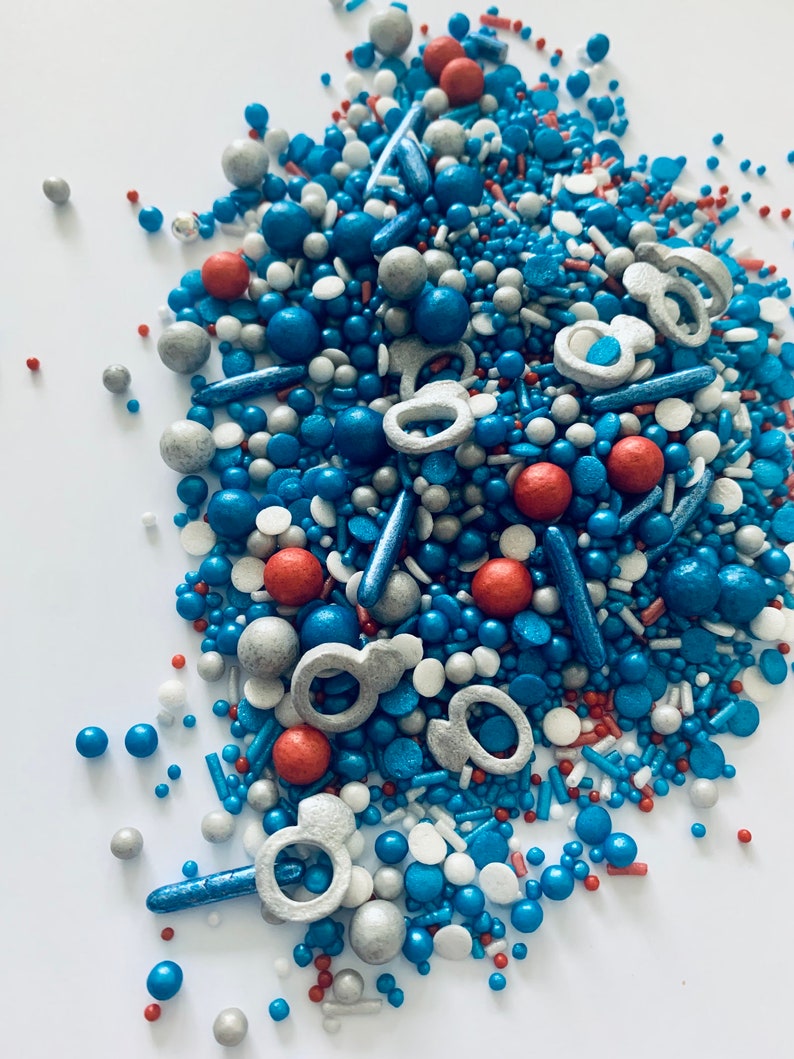 May include: A mix of blue, red, silver, and white sprinkles. The sprinkles are in the shape of balls, stars, and rings.