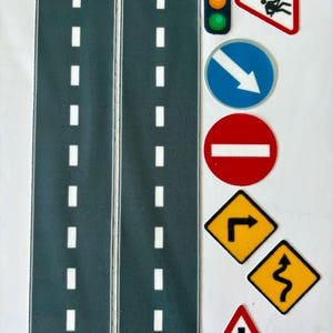 May include: A set of toy road pieces and traffic signs. The road pieces are dark gray with white dashed lines. Traffic signs include a stoplight, a yield sign, a do not enter sign, and several warning signs.