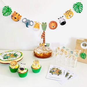 May include: A jungle-themed birthday party setup with a cake, cupcakes, and lollipops. The cake is decorated with a jungle scene and the number one. The cupcakes are topped with animal figurines. The lollipops are wrapped in clear cellophane and have animal designs. The party decorations include a banner with animal faces and green leaves.