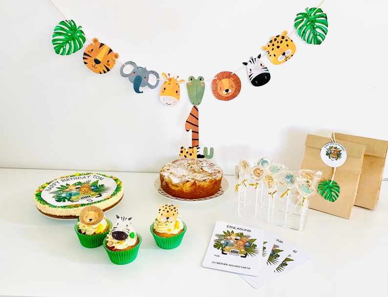 May include: A jungle-themed birthday party setup with a cake, cupcakes, and lollipops. The cake is decorated with a jungle scene and the number one. The cupcakes are topped with animal figurines. The lollipops are wrapped in clear cellophane and have animal designs. The party decorations include a banner with animal faces and green leaves.