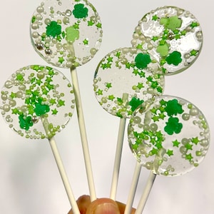 Puede incluir: Chupetines transparentes con decoraciones de tréboles y estrellas verdes. Los chupetines están en palitos blancos y tienen chispas plateadas y blancas. Se sostienen en una mano sobre un fondo blanco.