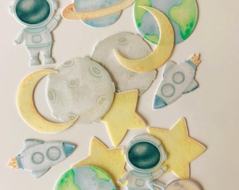 May include: A collection of space-themed paper cutouts featuring two astronauts in white suits, two rockets, two planets with rings, two Earth-like globes, two moons, and three yellow stars, all rendered in a soft watercolor style with pastel tones.