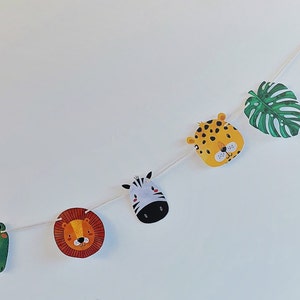 May include: A white string garland with green, yellow, orange, and black animal faces and a green leaf. The animals include a frog, lion, zebra, and leopard.