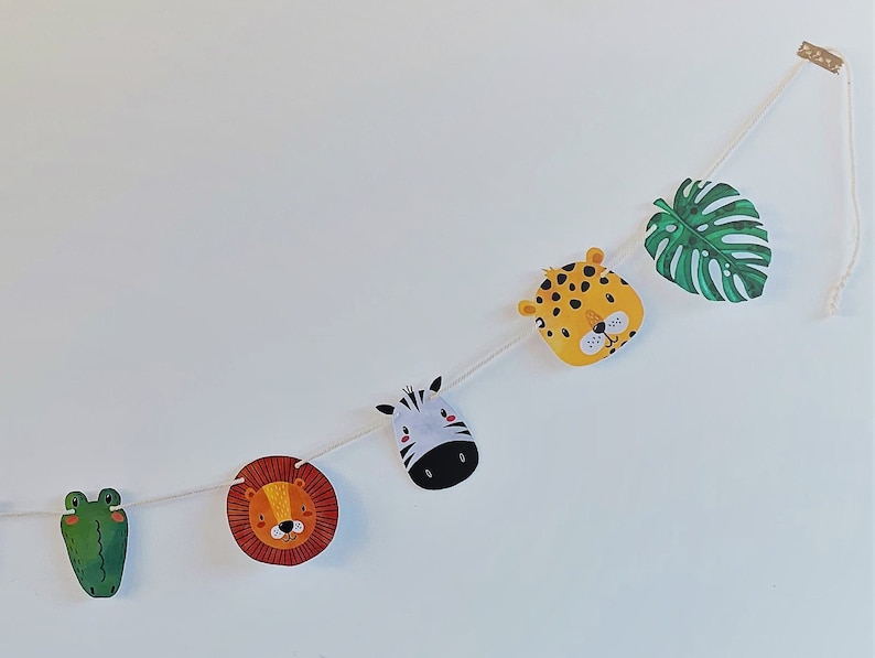 May include: A white string garland with green, yellow, orange, and black animal faces and a green leaf. The animals include a frog, lion, zebra, and leopard.