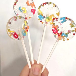 Puede incluir: Cuatro piruletas transparentes con letras coloridas y chispas. Cada piruleta tiene un palito blanco y un diseño de caramelo circular. Las letras son de varios colores, incluyendo rojo, azul, amarillo y naranja. Las chispas son pequeñas y multicolores.