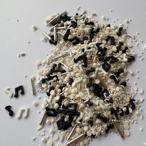 Puede incluir: Una mezcla de chispas negras, blancas y plateadas, que incluyen notas musicales, varillas y formas redondas. Las chispas están esparcidas sobre una superficie blanca, creando una textura visualmente atractiva. Ideal para decorar pasteles y otros productos horneados.