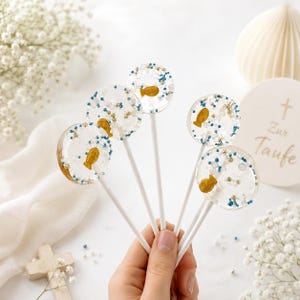 Può includere: Cinque lecca-lecca rotondi e trasparenti su bastoncini bianchi, contenenti ciascuno una piccola caramella a forma di pesce dorato, granelli blu e perline bianche. I lecca-lecca sono disposti a ventaglio, mettendo in mostra le decorazioni.