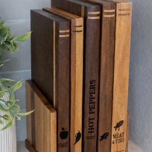 May include: A set of wooden cutting boards in a wooden holder. The boards are various shades of brown and feature etched designs and text, including an apple, a chili pepper, and the words "HOT PEPPERS" and "MEAT & FISH".