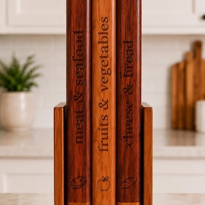 Puede incluir: Tres tablas de cortar de madera dispuestas verticalmente en un soporte de madera. Las tablas están grabadas con los textos meat & seafood, fruits & vegetables y cheese & bread, cada una con un pequeño icono de un filete, una manzana y un pan.