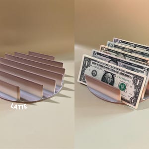 Puede incluir: Un organizador de efectivo de metal color oro rosa con un dise&ntilde;o curvo y m&uacute;ltiples ranuras para guardar billetes. El organizador est&aacute; lleno de billetes de d&oacute;lar estadounidense, incluyendo un billete de 1$, 5$, 10$, 20$ y 50$.