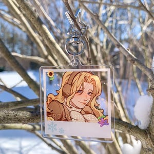 May include: A clear acrylic keychain featuring a cartoon illustration of a person with blonde hair wearing headphones. The illustration is in a polaroid-style frame with a sunflower, snowflake, and flower accents. The keychain is attached to a silver star-shaped clasp.