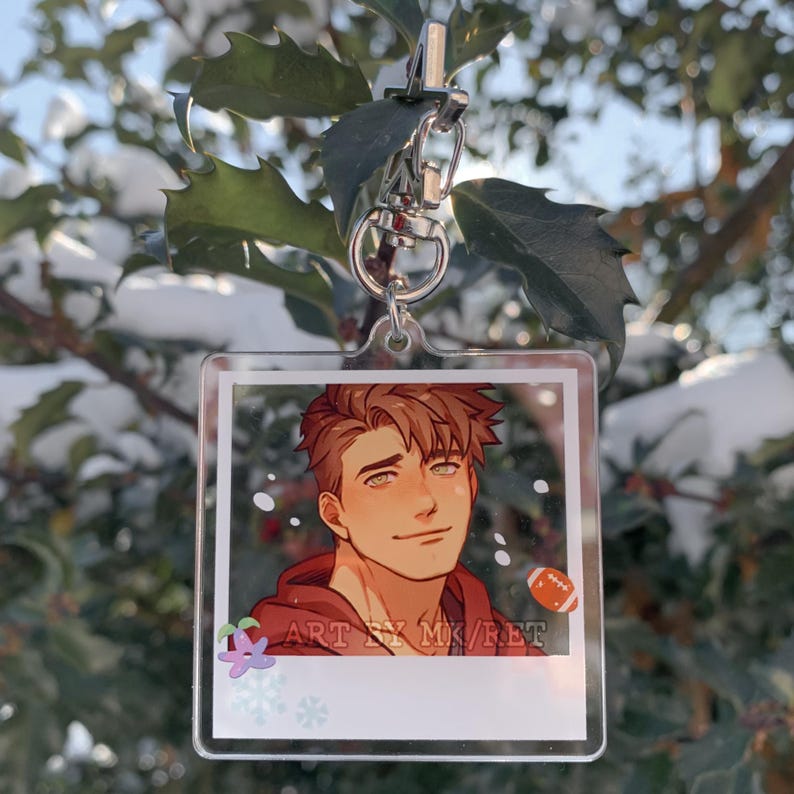 May include: A clear acrylic keychain featuring a cartoon illustration of a person with brown hair and a red hoodie. The keychain has a silver clasp and a small football graphic. Text on the keychain reads "ART BY MK/RET".