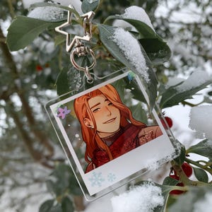 May include: A clear acrylic keychain featuring a smiling character with long red hair. The keychain has a silver star-shaped clasp and a small illustration of a flower. The character is wearing a red scarf with text that reads "BY MK RET".