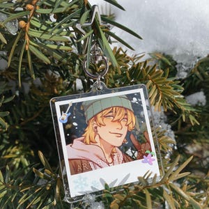 May include: A clear acrylic keychain featuring a cartoon illustration of a person with blonde hair, wearing a green beanie and a pink hoodie. The keychain has a silver clasp and is hanging from a tree branch. The text "ART BY MK" is visible.