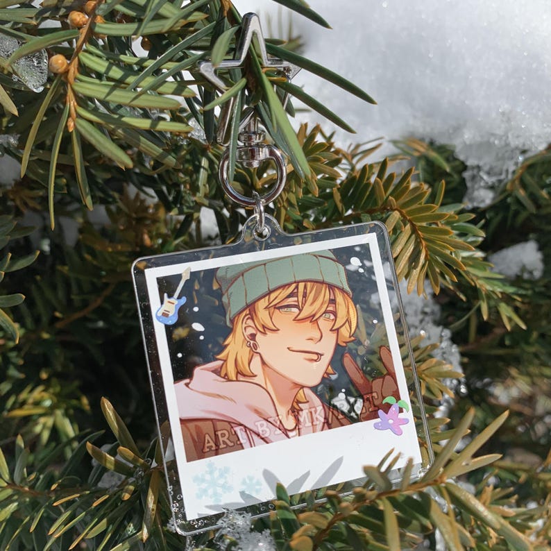 May include: A clear acrylic keychain featuring a cartoon illustration of a person with blonde hair, wearing a green beanie and a pink hoodie. The keychain has a silver clasp and is hanging from a tree branch. The text "ART BY MK" is visible.