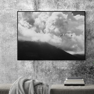 Puede incluir: Fotograf&iacute;a en blanco y negro enmarcada de un paisaje monta&ntilde;oso con un cielo nublado. Una manta gris est&aacute; sobre un otom&aacute;n gris oscuro. Una pila de libros est&aacute; sobre un otom&aacute;n a juego. La pared es gris texturizada.