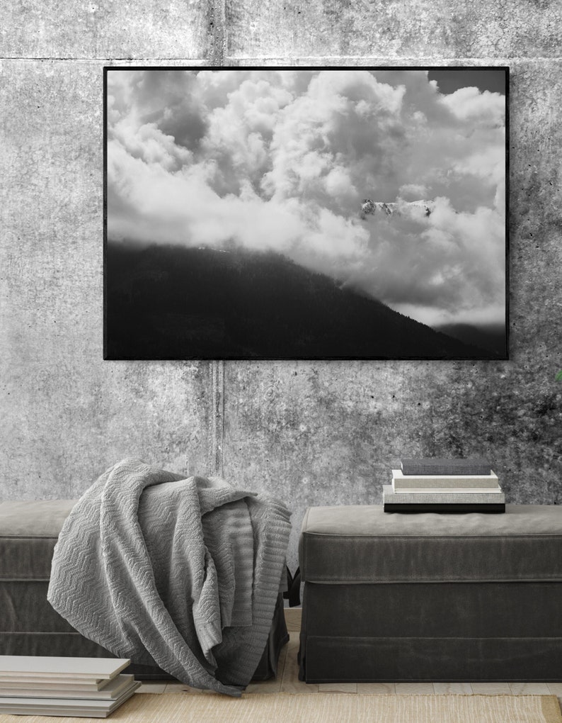 Puede incluir: Fotograf&iacute;a en blanco y negro enmarcada de un paisaje monta&ntilde;oso con un cielo nublado. Una manta gris est&aacute; sobre un otom&aacute;n gris oscuro. Una pila de libros est&aacute; sobre un otom&aacute;n a juego. La pared es gris texturizada.