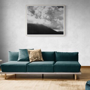 Puede incluir: Una fotograf&iacute;a en blanco y negro enmarcada de una cordillera nublada cuelga sobre un sof&aacute; seccional verde azulado. El sof&aacute; tiene varias almohadas, incluida una dorada con lentejuelas. A la izquierda hay una peque&ntilde;a mesa auxiliar con un jarr&oacute;n de cristal.