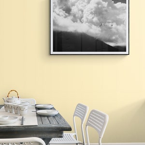 Puede incluir: Fotograf&iacute;a en blanco y negro enmarcada de una monta&ntilde;a parcialmente oscurecida por grandes y esponjosas nubes. La fotograf&iacute;a cuelga de una pared amarillo p&aacute;lido sobre una mesa de comedor con vajilla y sillas blancas.