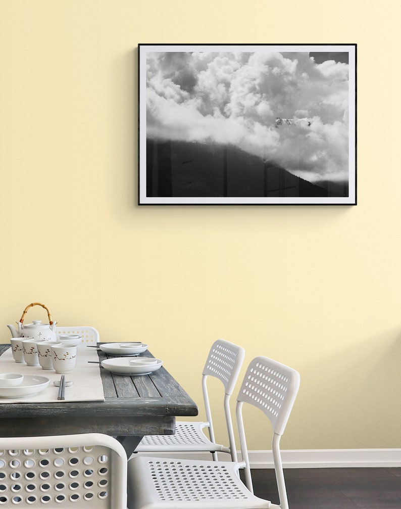 Puede incluir: Fotograf&iacute;a en blanco y negro enmarcada de una monta&ntilde;a parcialmente oscurecida por grandes y esponjosas nubes. La fotograf&iacute;a cuelga de una pared amarillo p&aacute;lido sobre una mesa de comedor con vajilla y sillas blancas.
