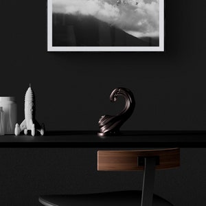 Puede incluir: Una impresi&oacute;n enmarcada en blanco y negro de una escena de monta&ntilde;a con nubes cuelga sobre una estanter&iacute;a negra. En la estanter&iacute;a hay una figura de cohete blanca y una escultura de onda de bronce oscuro. Una silla negra con respaldo de madera est&aacute; en primer plano.