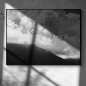 Puede incluir: Fotograf&iacute;a en blanco y negro enmarcada de un paisaje monta&ntilde;oso con nubes. La imagen captura una cordillera con un bosque en la base, parcialmente oculta por las nubes. El cielo est&aacute; lleno de nubes oscuras y dram&aacute;ticas, iluminadas por la luz del sol.