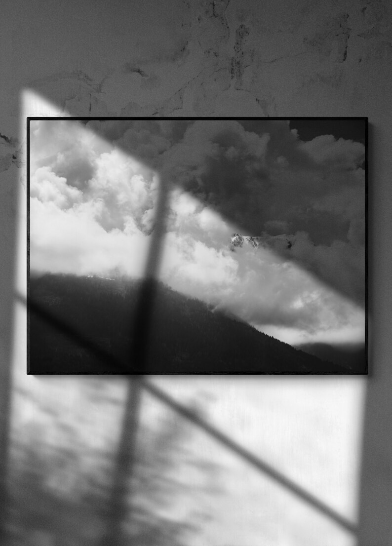 Puede incluir: Fotograf&iacute;a en blanco y negro enmarcada de un paisaje monta&ntilde;oso con nubes. La imagen captura una cordillera con un bosque en la base, parcialmente oculta por las nubes. El cielo est&aacute; lleno de nubes oscuras y dram&aacute;ticas, iluminadas por la luz del sol.