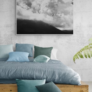 Puede incluir: Una escena de dormitorio con una cama con un edred&oacute;n azul gris&aacute;ceo y varias almohadas azules y verdes. Una fotograf&iacute;a en blanco y negro de un paisaje de monta&ntilde;a y nubes cuelga sobre la cama. Una plataforma de madera sirve como estructura de la cama.