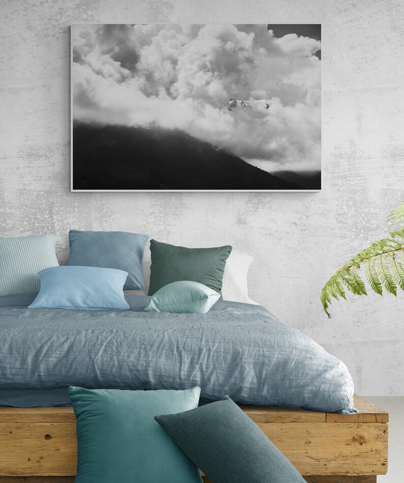 Puede incluir: Una escena de dormitorio con una cama con un edred&oacute;n azul gris&aacute;ceo y varias almohadas azules y verdes. Una fotograf&iacute;a en blanco y negro de un paisaje de monta&ntilde;a y nubes cuelga sobre la cama. Una plataforma de madera sirve como estructura de la cama.