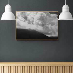 Puede incluir: Impresi&oacute;n enmarcada en blanco y negro de un paisaje monta&ntilde;oso con nubes dram&aacute;ticas. La obra de arte est&aacute; colgada en una pared gris oscuro, iluminada por dos l&aacute;mparas colgantes blancas. Debajo hay una superficie de madera con listones.
