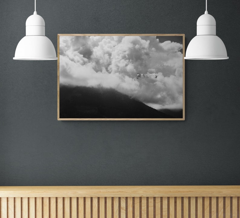 Puede incluir: Impresi&oacute;n enmarcada en blanco y negro de un paisaje monta&ntilde;oso con nubes dram&aacute;ticas. La obra de arte est&aacute; colgada en una pared gris oscuro, iluminada por dos l&aacute;mparas colgantes blancas. Debajo hay una superficie de madera con listones.