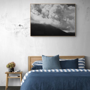 Puede incluir: Una fotograf&iacute;a en blanco y negro enmarcada de una cordillera nublada cuelga sobre una cama. La cama presenta un cabecero de madera, ropa de cama azul y almohadas decorativas. Una peque&ntilde;a mesita de noche de madera con una planta y decoraci&oacute;n est&aacute; junto a la cama.