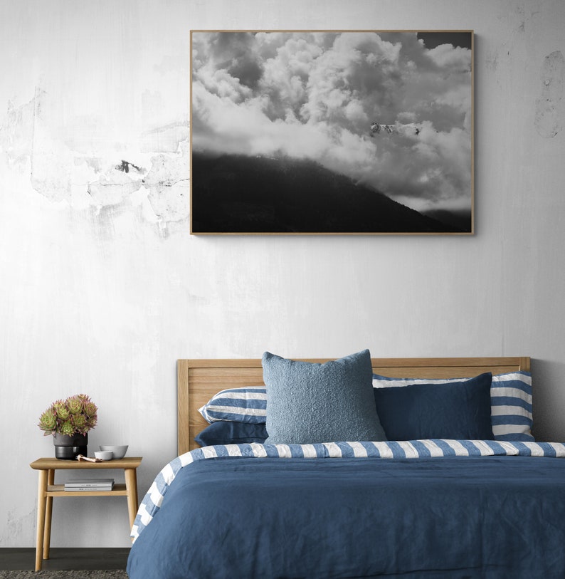 Puede incluir: Una fotograf&iacute;a en blanco y negro enmarcada de una cordillera nublada cuelga sobre una cama. La cama presenta un cabecero de madera, ropa de cama azul y almohadas decorativas. Una peque&ntilde;a mesita de noche de madera con una planta y decoraci&oacute;n est&aacute; junto a la cama.