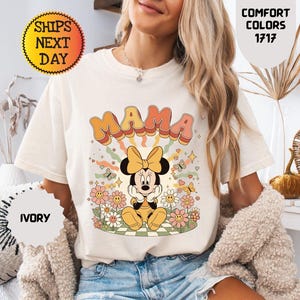 May include: Ivory-colored t-shirt featuring the word "MAMA" in retro orange and pink lettering, with a Minnie Mouse graphic. The design includes flowers, butterflies, and a yellow bow. The shirt is part of the Comfort Colors 1717 collection.