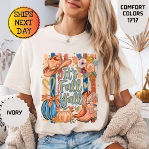 May include: An ivory short-sleeve t-shirt featuring a graphic print with a cowboy hat, floral arrangements, a blue pumpkin, an orange pumpkin, and cowboy boots. The text reads It's Fall Y'all in blue script. The shirt is a Comfort Colors 1717 model.