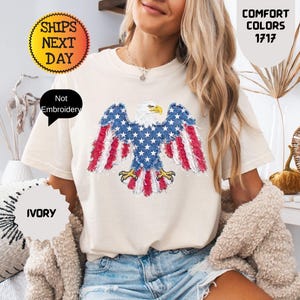 May include: Ivory-colored t-shirt featuring a patriotic eagle design with red, white, and blue accents. The shirt has short sleeves and a crew neck. The text "Comfort Colors 1717" is visible. The shirt is paired with denim shorts.