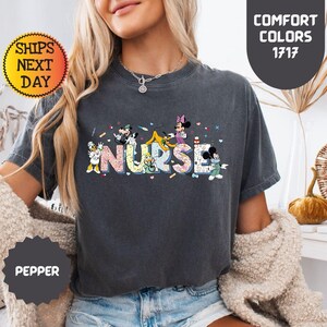 May include: A dark gray t-shirt with the word "NURSE" in colorful letters, featuring cartoon characters. The shirt has a round neck and short sleeves. The text "COMFORT COLORS 1717" is on the upper right.