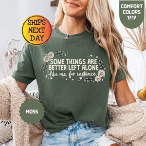May include: Moss green t-shirt with the text "SOME THINGS ARE BETTER LEFT ALONE like me, for instance." The shirt has a floral design and is part of the Comfort Colors collection. The image also includes a "Ships Next Day" sticker.