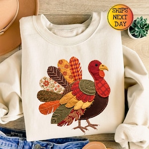 Könnte beinhalten: Cremefarbenes Sweatshirt mit einem bunten Truthahndesign. Der Truthahn hat einen roten Kopf, einen gelben Schnabel und einen Patchwork aus gemusterten Stoffen für seinen Körper und seine Federn. Der Text "SHIPS NEXT DAY" befindet sich in einem gelben Kreis.