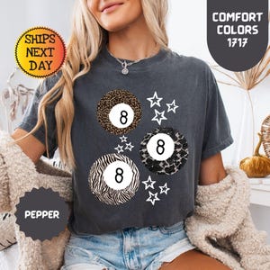 May include: Dark gray t-shirt featuring three stylized 8-ball designs with animal print patterns and white star accents. The shirt has the text "COMFORT COLORS 1717" and "PEPPER".