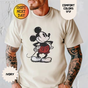 May include: Ivory-colored t-shirt featuring a sketch-style Mickey Mouse graphic in black, red, and white. The shirt has the text "Comfort Colors 1717" and "Ships Next Day".