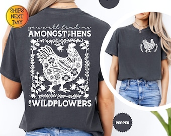 以下が含まれることがあります： ホワイトのグラフィックプリントが施されたチャコールグレーのTシャツ。背面には野花に囲まれた大きな雌鶏と「you will find me AMONGST THE HENS AND WILDFLOWERS」の文字が描かれています。胸元には小さめの雌鶏のグラフィックがあります。