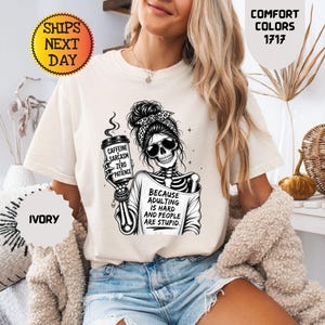 以下が含まれることがあります： アイボリーの半袖Tシャツ。お団子ヘア、サングラス、コーヒーカップを持つ骸骨の黒いグラフィックが描かれています。カップには「CAFFEINE SARCASM ZERO PATIENCE」、Tシャツには「BECAUSE ADULTING IS HARD AND PEOPLE ARE STUPID」と書かれています。