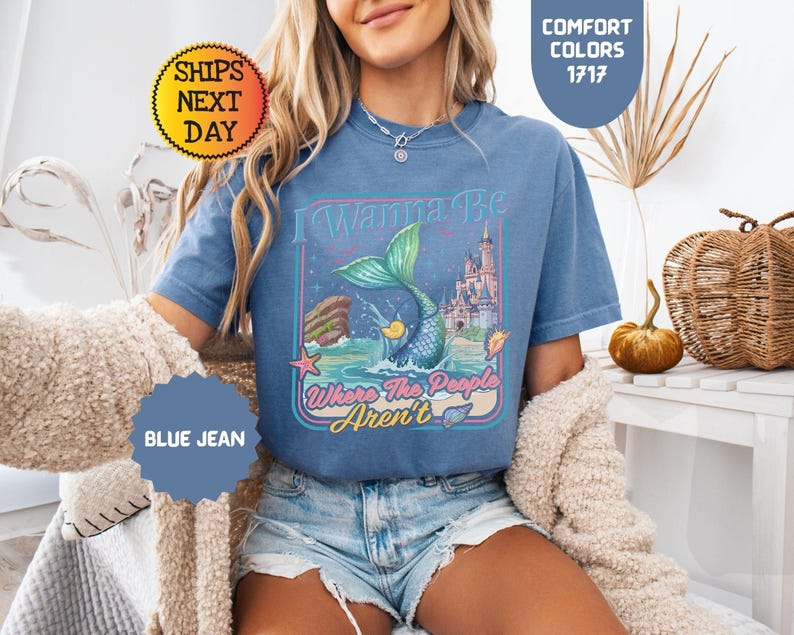 May include: A blue jean colored t-shirt featuring a graphic print of a mermaid tail splashing in the ocean, a castle, and the text I Wanna Be Where The People Aren't. The shirt has a crew neckline and short sleeves, showcasing a vintage-inspired design.