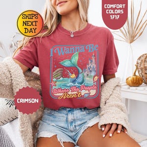 May include: A crimson short-sleeve t-shirt featuring a graphic print of a mermaid tail emerging from the ocean, a castle, and the text I Wanna Be Where The People Aren't. The design includes a starfish, a seashell, and a small yellow duck.