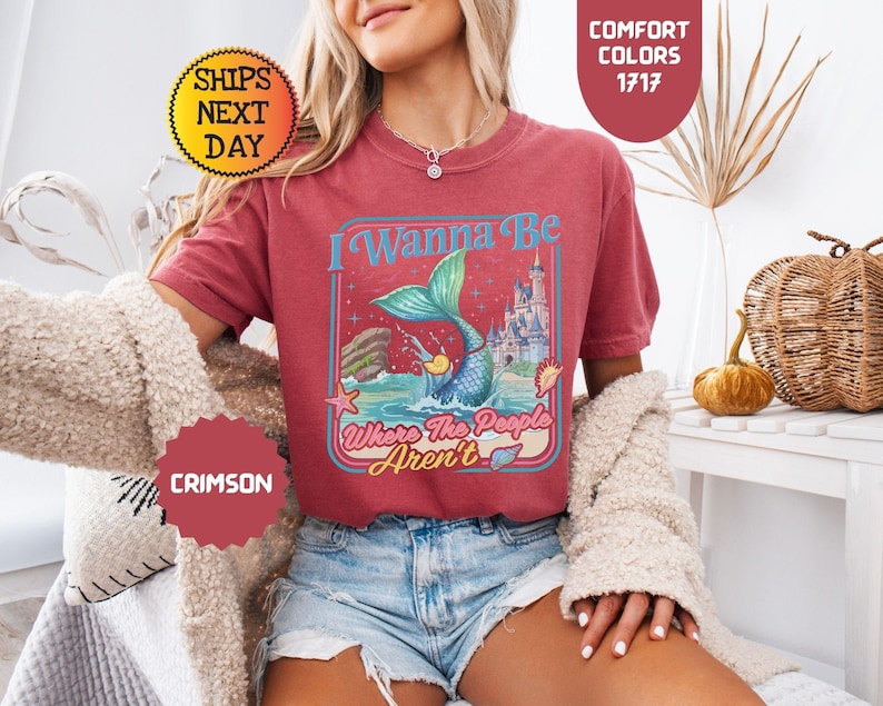 May include: A crimson short-sleeve t-shirt featuring a graphic print of a mermaid tail emerging from the ocean, a castle, and the text I Wanna Be Where The People Aren't. The design includes a starfish, a seashell, and a small yellow duck.